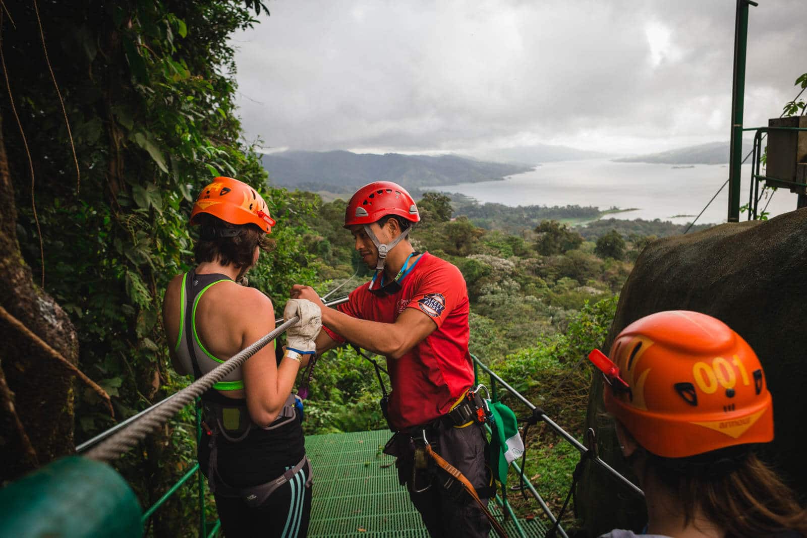 Un guía ayuda a una mujer a prepararse para hacer tirolesa en una plataforma con vista al lago Arenal y la selva